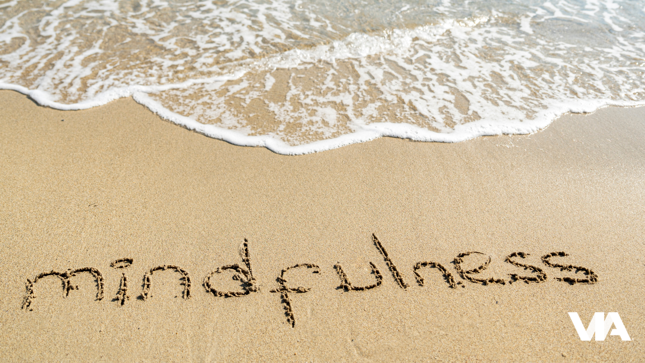 The word ‘mindfulness’ written in the sand beside gentle waves, evoking calm and presence as the water approaches.