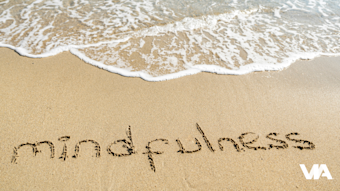The word ‘mindfulness’ written in the sand beside gentle waves, evoking calm and presence as the water approaches.